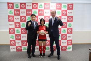 中央でラグビーボールを手にとり写真に写る松田さん。両脇にはこぶしを握った武井市長と桑原教育長